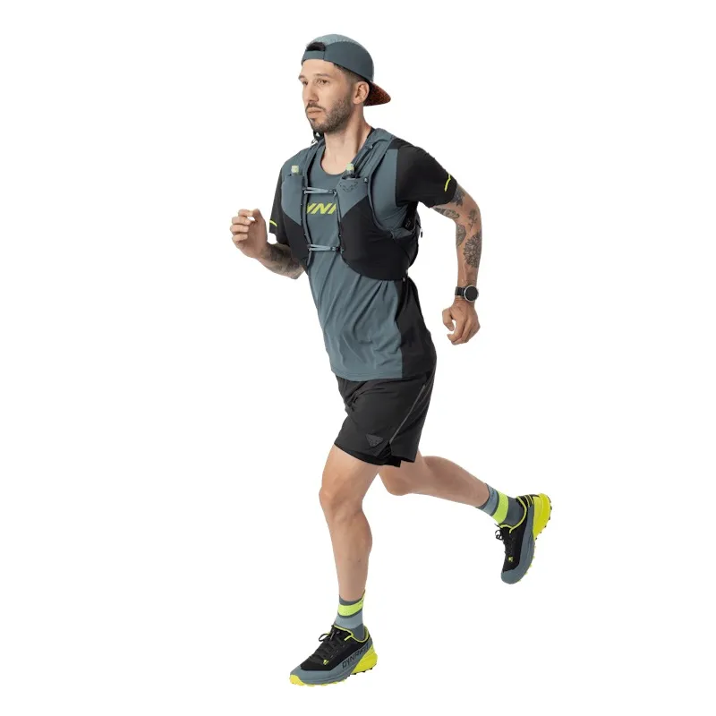 Ein Mann mit Bart, Kappe und Laufweste rennt vor einem weißen Hintergrund. Er trägt ein graues Laufshirt, schwarze Shorts, Sportschuhe und eine Uhr. Auf seinem linken Arm befindet sich ein Tattoo.