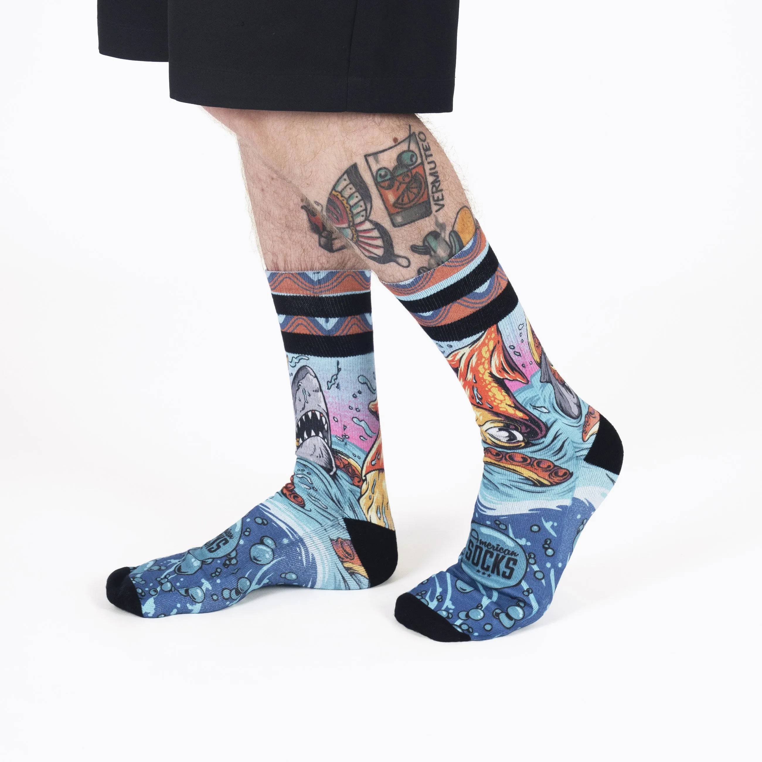 Zwei Beine mit tätowierten Waden tragen blaue `Seamonsters` Socken mit Hai, Oktopus und Blasen Motiven.