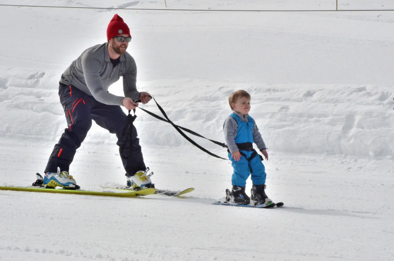 Vater zieht Kind mit SkiTies Ultimate Kid's Ski Harness auf Skiern im Schnee.
