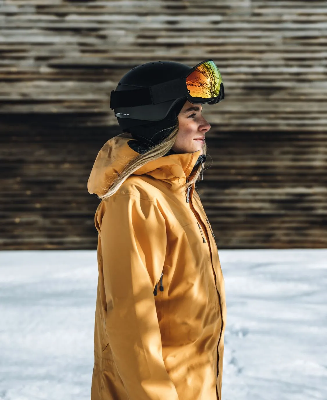 Profilansicht einer Person mit schwarzem POC Meninx Skihelm und gelber Jacke im Schnee. Die Skibrille spiegelt Bäume wider.
