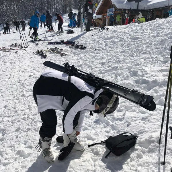 Skifahrerin beugt sich vor, um Skischuhe zu befestigen, Ski mit Skiback auf dem Rücken. Verschneite Landschaft.