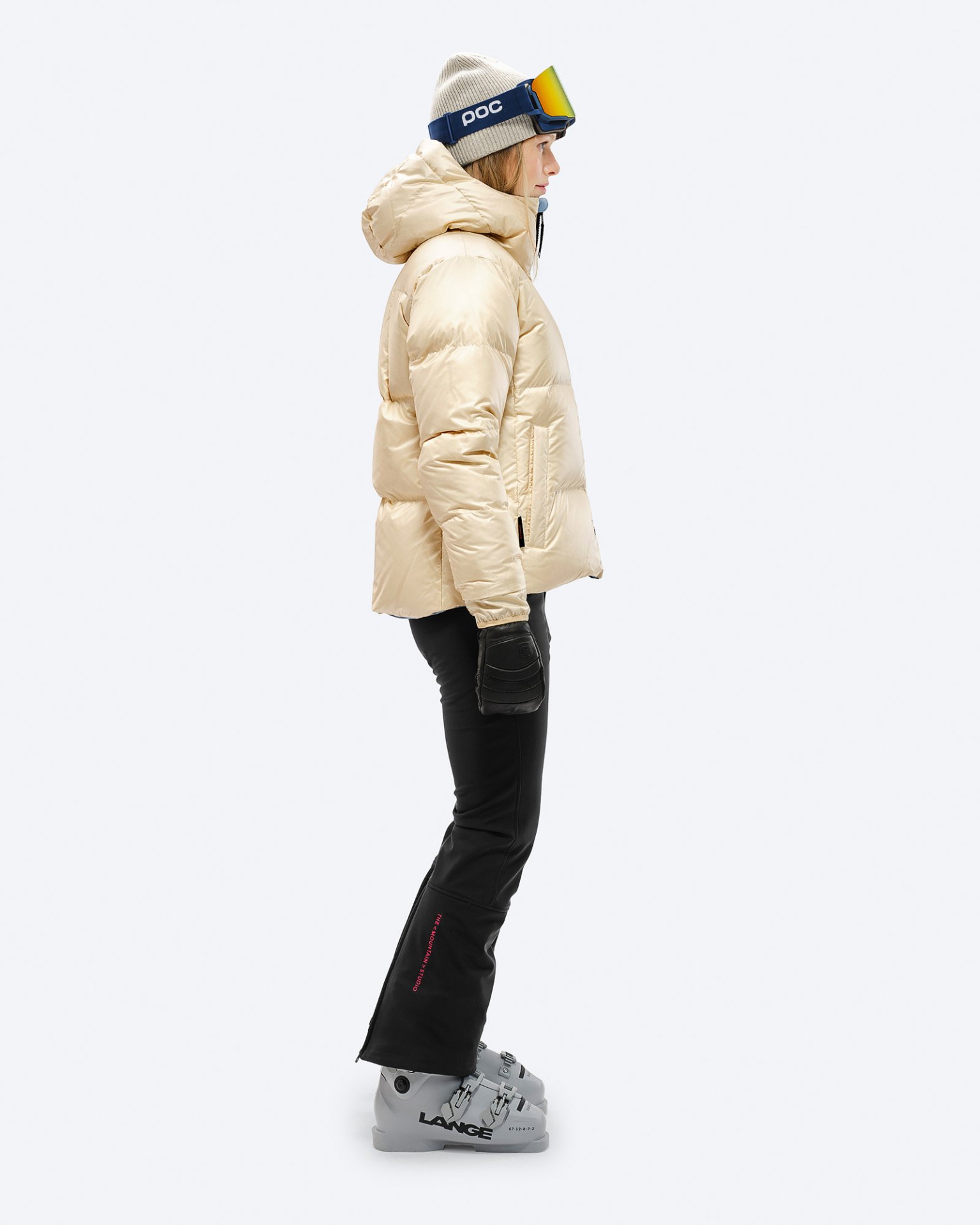 Frau in beiger Daunenjacke mit Kapuze, Skihose und Skischuhen in Seitenansicht. Winterbekleidung.