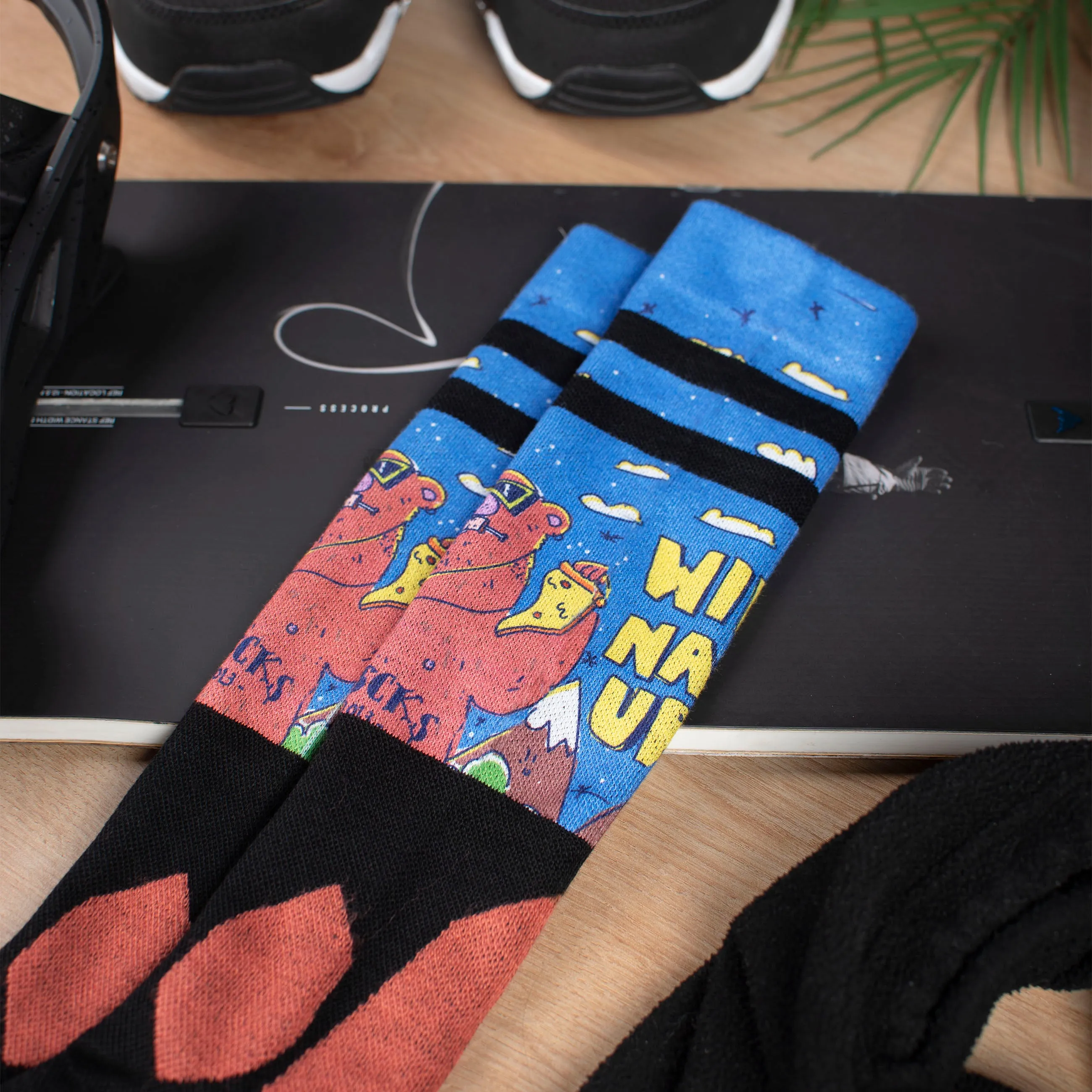 Snow Socks mit Bärenmotiv liegen auf einem Snowboard, Boots und Grün im Hintergrund.