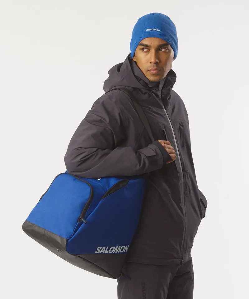 Mann mit blauer Mütze, trägt Salomon Skischuhtasche über der Schulter. Die Tasche ist blau-schwarz mit Salomon Logo.