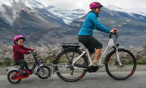 Frau und Kind mit FollowMe Tandemkupplung auf Fahrrädern vor Berglandschaft.