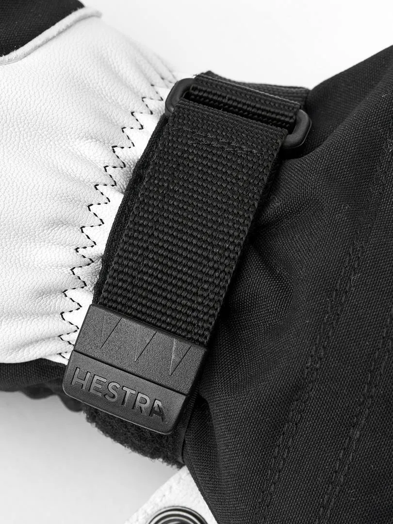 Detailaufnahme eines schwarzen Hestra Skihandschuhs mit weißem Lederteil und einem schwarzen Gurt mit dem Hestra-Logo.