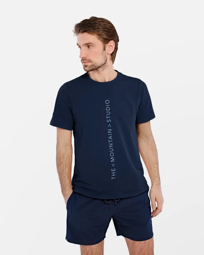 Mann trägt dunkelblaues T-Shirt mit vertikalem "The Mountain Studio"-Logo und passende Shorts.