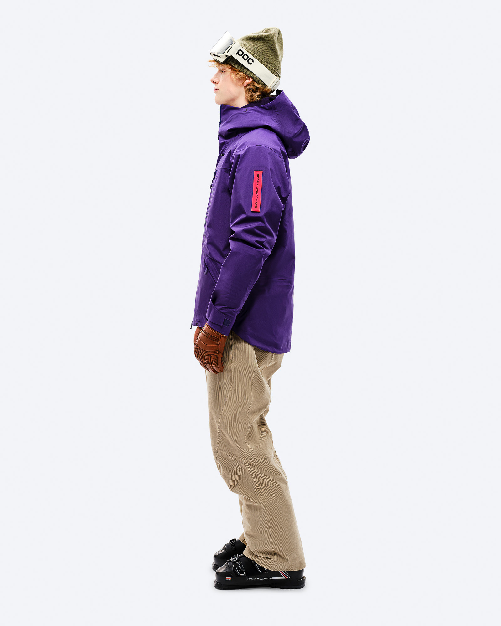 Person in lila GORE-TEX EPE 3L Shell Jacket, Skihose, Handschuhe und Skischuhe, seitliche Ansicht.