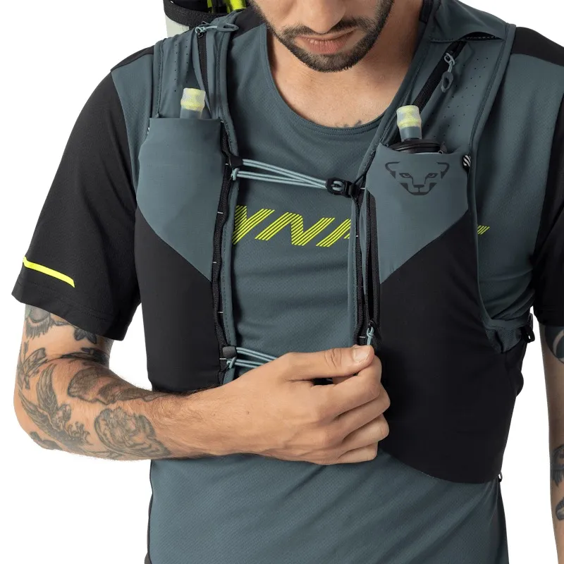 Ein Mann mit Bart trägt eine Laufweste mit Trinkflaschen in den Taschen. Er öffnet den Reißverschluss der Weste. Die Weste ist grau und schwarz. Er trägt ein graues Laufshirt mit gelbem Aufdruck und kurze, schwarze Ärmel. Sein Arm ist tätowiert.