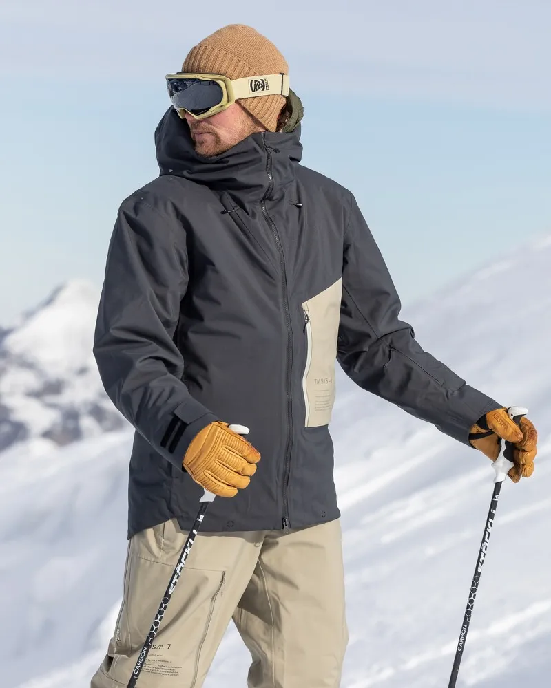 Mann mit Skibrille und Mütze trägt graue Ride Insulated Jacket und Handschuhe, hält Skistöcke im Schnee.
