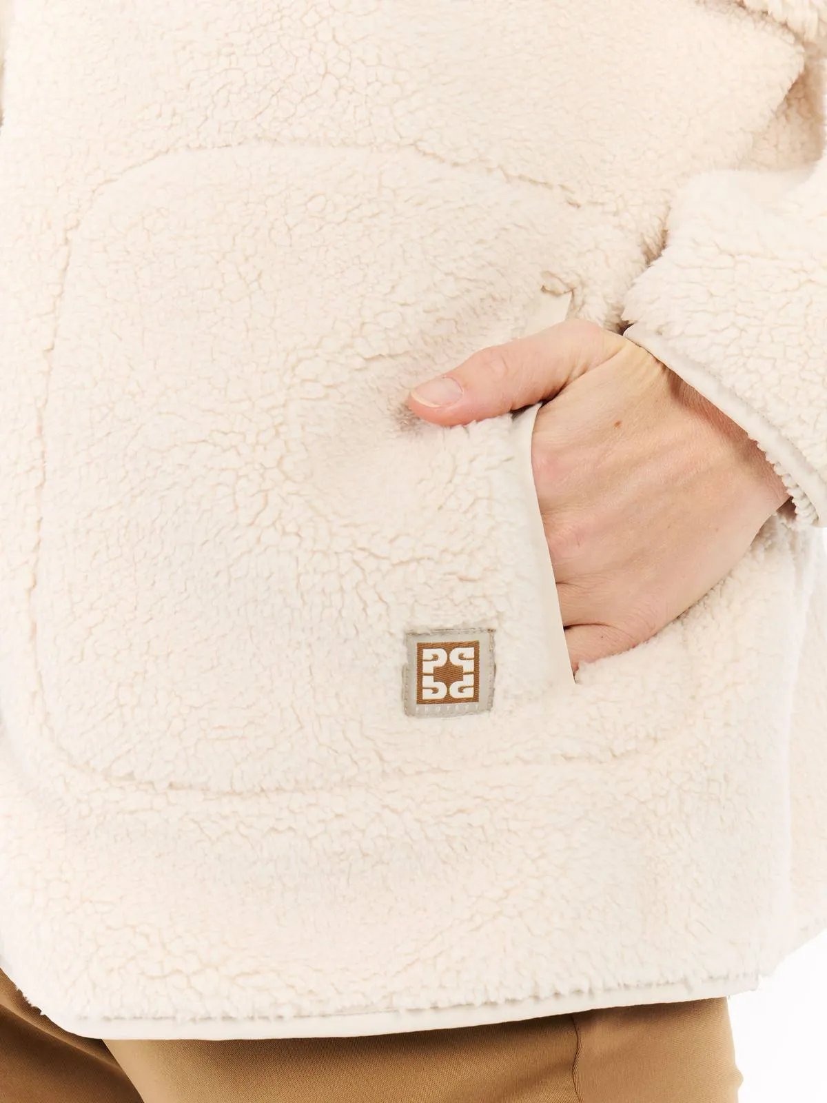 Nahaufnahme einer beigen Fleecejacke mit einer Hand in der Tasche und einem kleinen, quadratischen Logo auf der linken Seite.