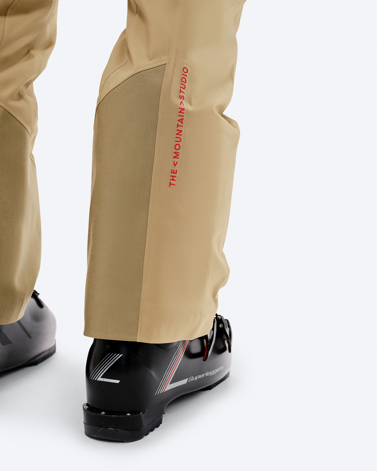 Detailaufnahme der P-1 GORE-TEX Hose in Sandfarbe, die über Skischuhen getragen wird. Aufgedrucktes Markenlogo.