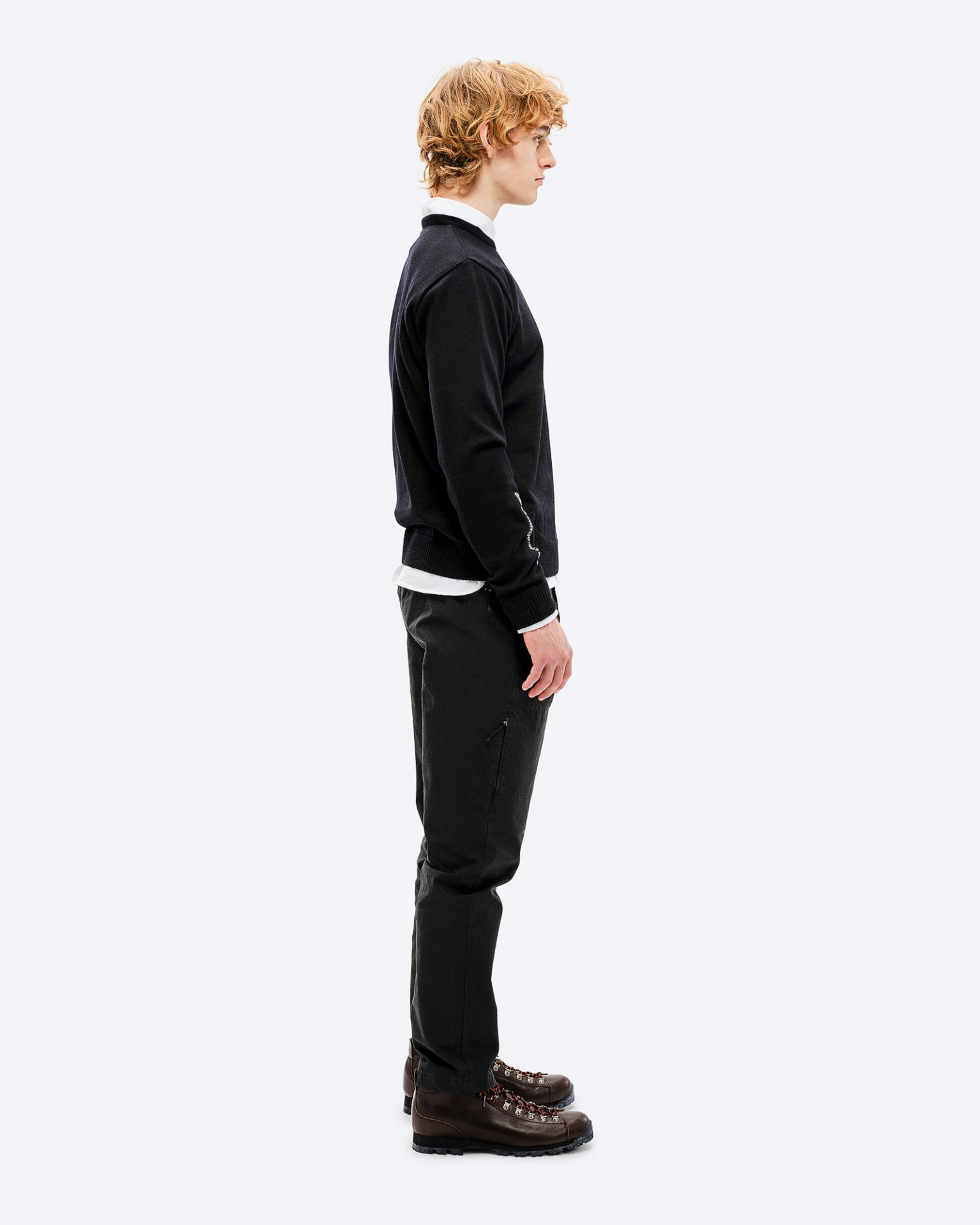 Ein Mann im Profil trägt einen schwarzen K-3 Ski Sweater aus Merinowolle, dazu dunkle Hosen und braune Wanderschuhe.