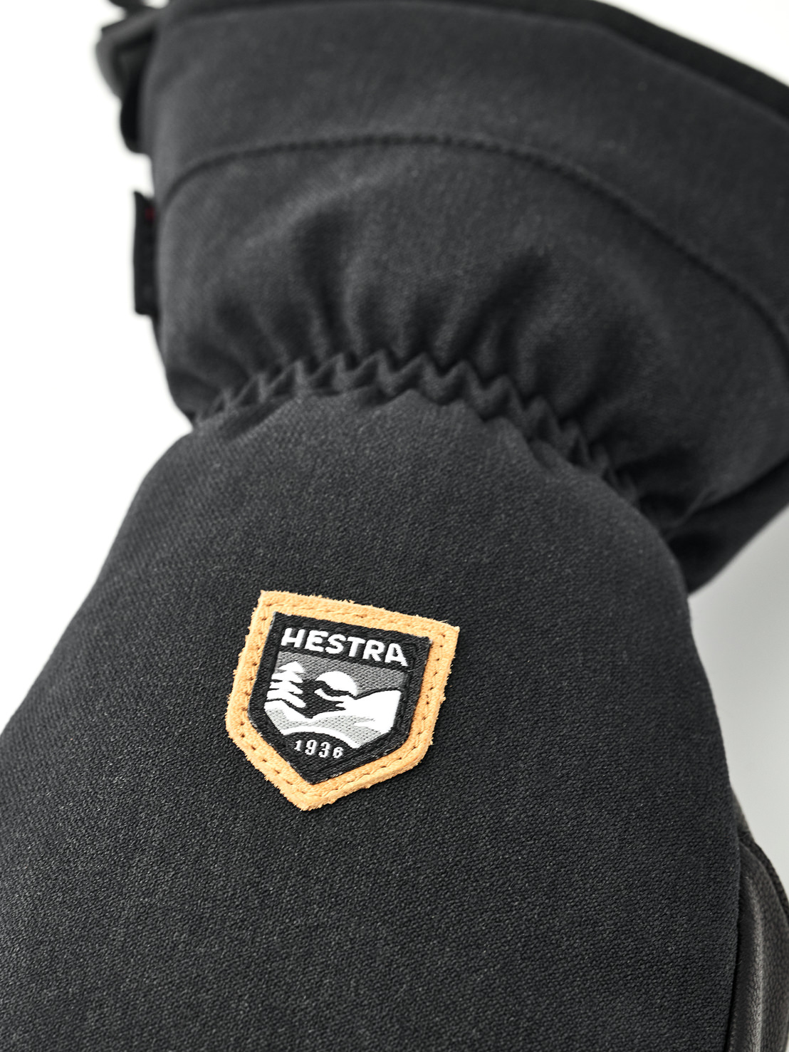Detailaufnahme eines schwarzen Fäustlings mit dem Hestra-Logo, einem orange umrandeten Schild mit Bergmotiv und dem Gründungsjahr 1936.