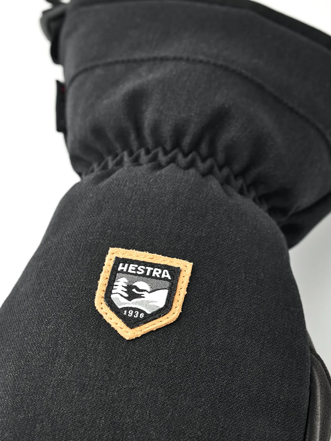 Detailaufnahme eines schwarzen Fäustlings mit dem Hestra-Logo, einem orange umrandeten Schild mit Bergmotiv und dem Gründungsjahr 1936.