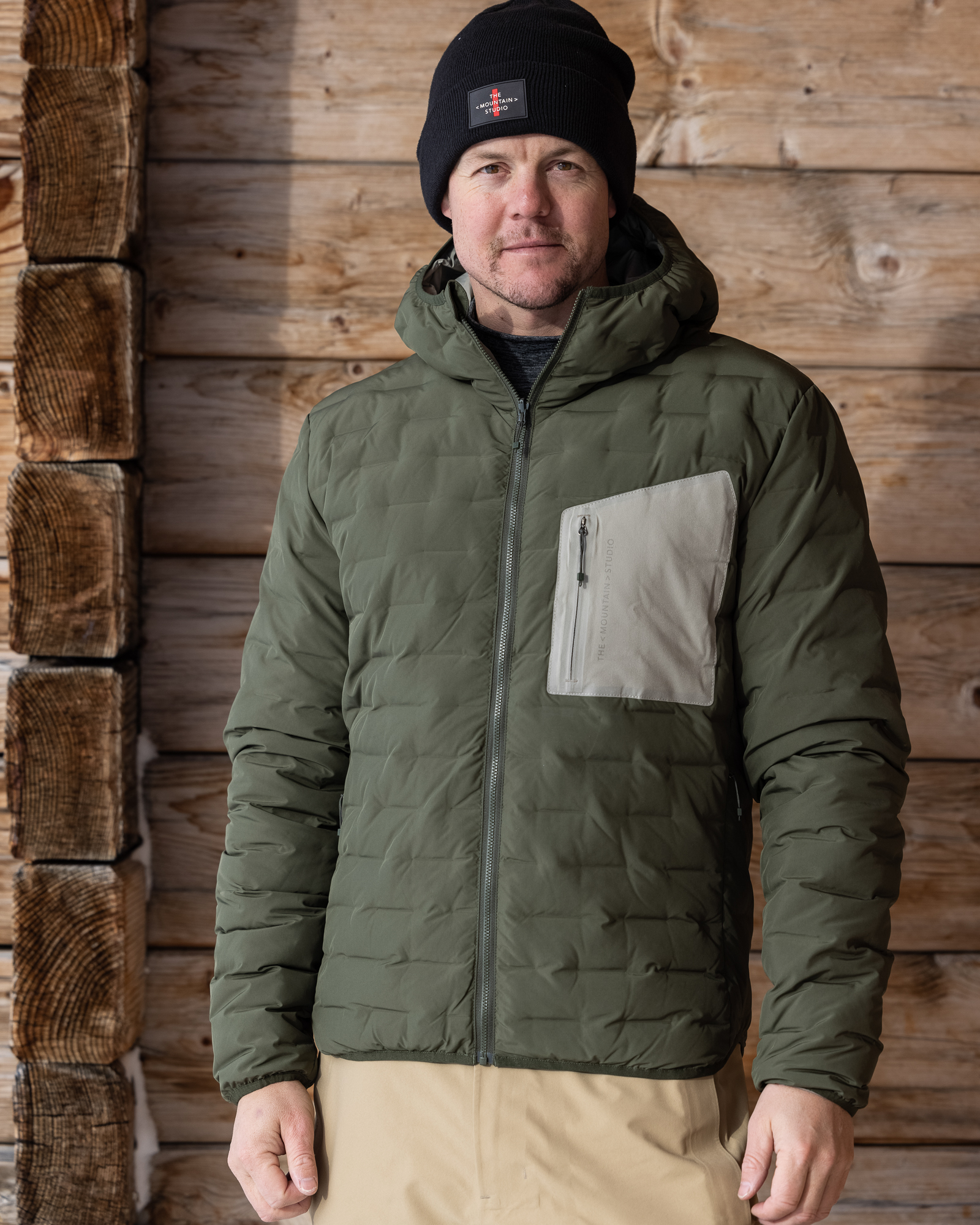 Ein Mann in D-3L Reversible Light Hood Jacket Forest Green mit Kapuze und Mütze vor einer Holzwand.