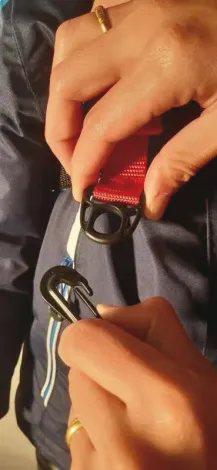 Detailaufnahme: Hände befestigen rote Leine mit Karabiner am Ski Trainer Geschirr. Fokus auf Verschlussmechanismus.