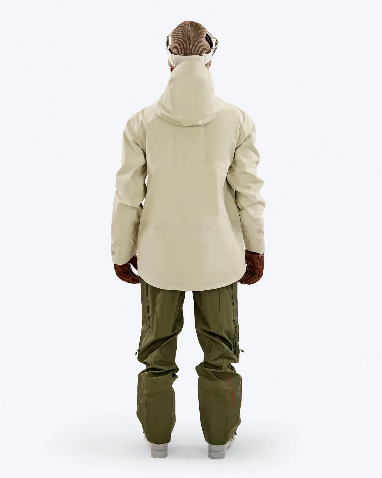 Rückansicht einer Person in Skikleidung: beige Jacke, grüne Hose, braune Handschuhe, Mütze und Skibrille auf weißem Hintergrund.