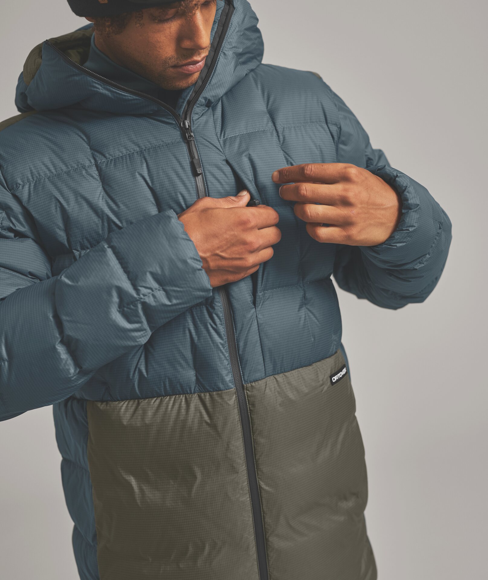 Kapuzen-Daunenjacke ORTOVOX Person schliesst die Kapuzen-Daunenjacke; obere Hälfte blau, untere olive; Markenzeichen "ORTVOX" auf Hüfthöhe.