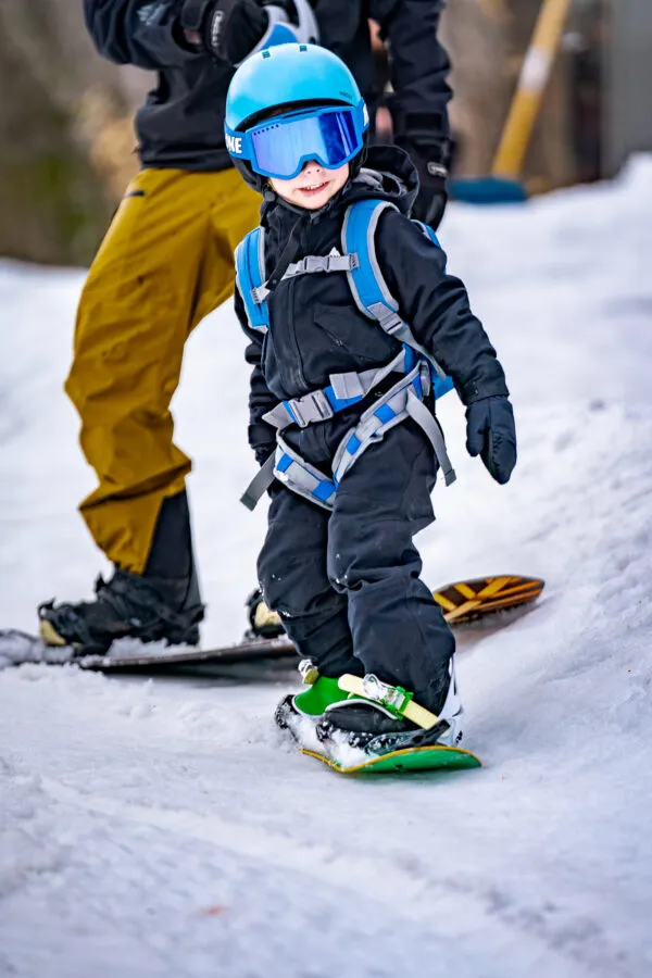 Kind mit blauem Helm und Ski-Brille, mit "The One" Snowboard Harness, lernt Snowboard fahren.