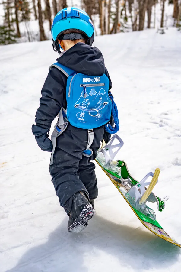 Kleines Kind mit blauem Helm, Rucksack und Snowboard auf einer verschneiten Piste, von hinten gesehen.