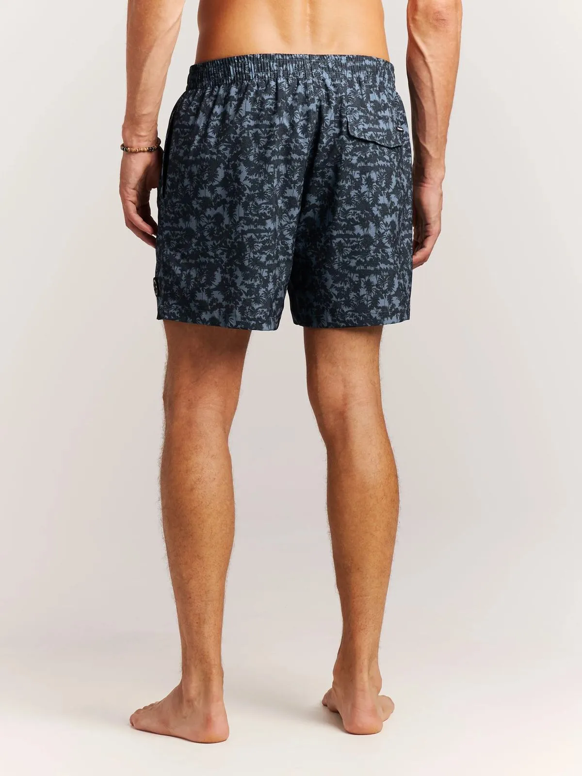 Ein Mann mit Badehose, von hinten gesehen, steht barfuß auf einem weißen Hintergrund. Seine Badehose ist blau mit dunklem Palmenmuster.