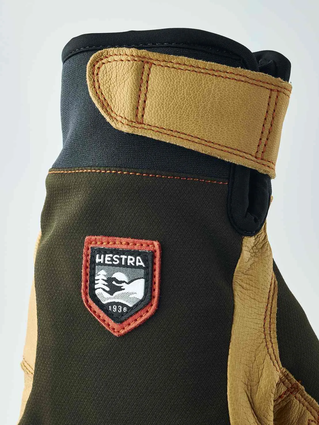 Detailaufnahme eines Handschuhs mit braunem Obermaterial, Lederdetails und "Hestra 1936" Logo. Der Handschuh hat eine Stulpe mit Klettverschluss.