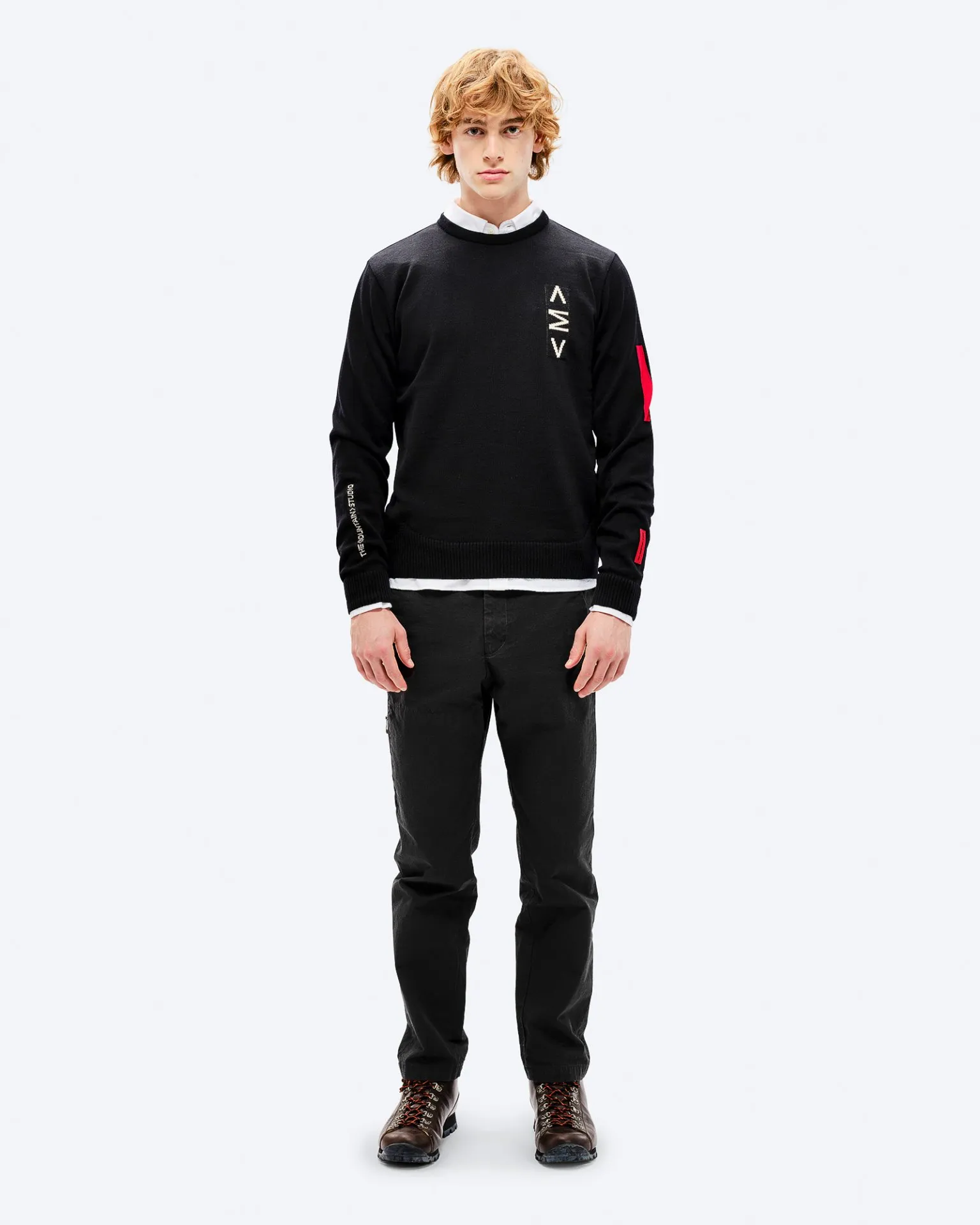 Mann mit rotem Haar trägt schwarzen K-3 Ski Sweater mit roten Akzenten und schwarzen Hosen, posiert vor einem weißen Hintergrund.