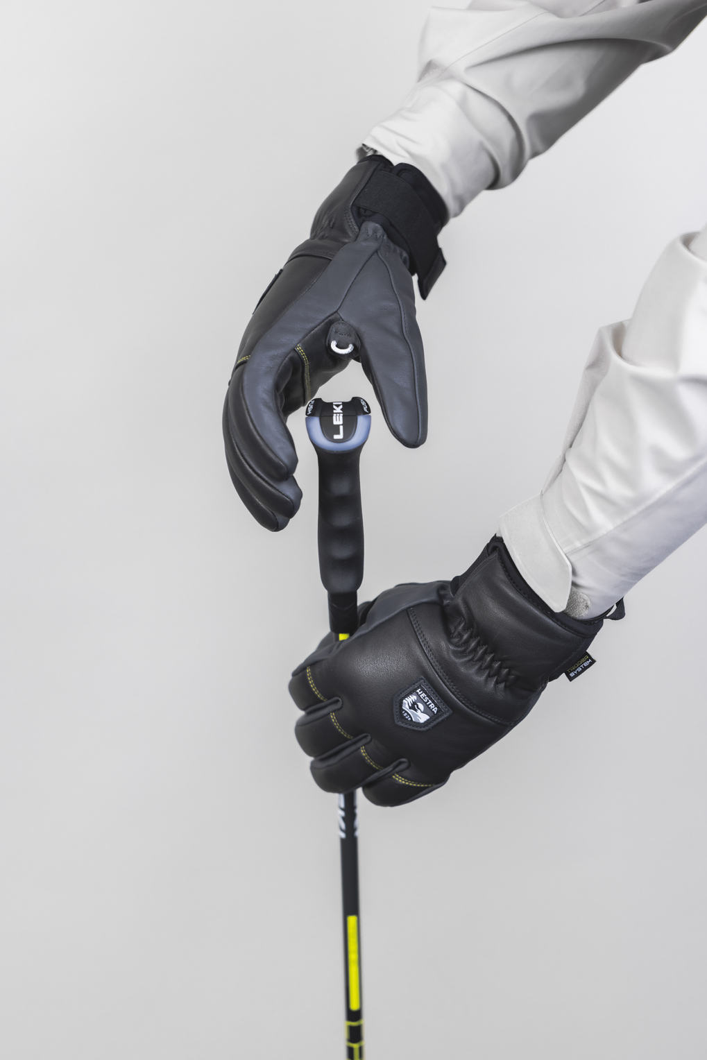 Hestra Omni Trigger jetzt Neu! Zwei behandschuhte Hände halten einen schwarzen Skistock mit gelben Akzenten vor einem hellgrauen Hintergrund.