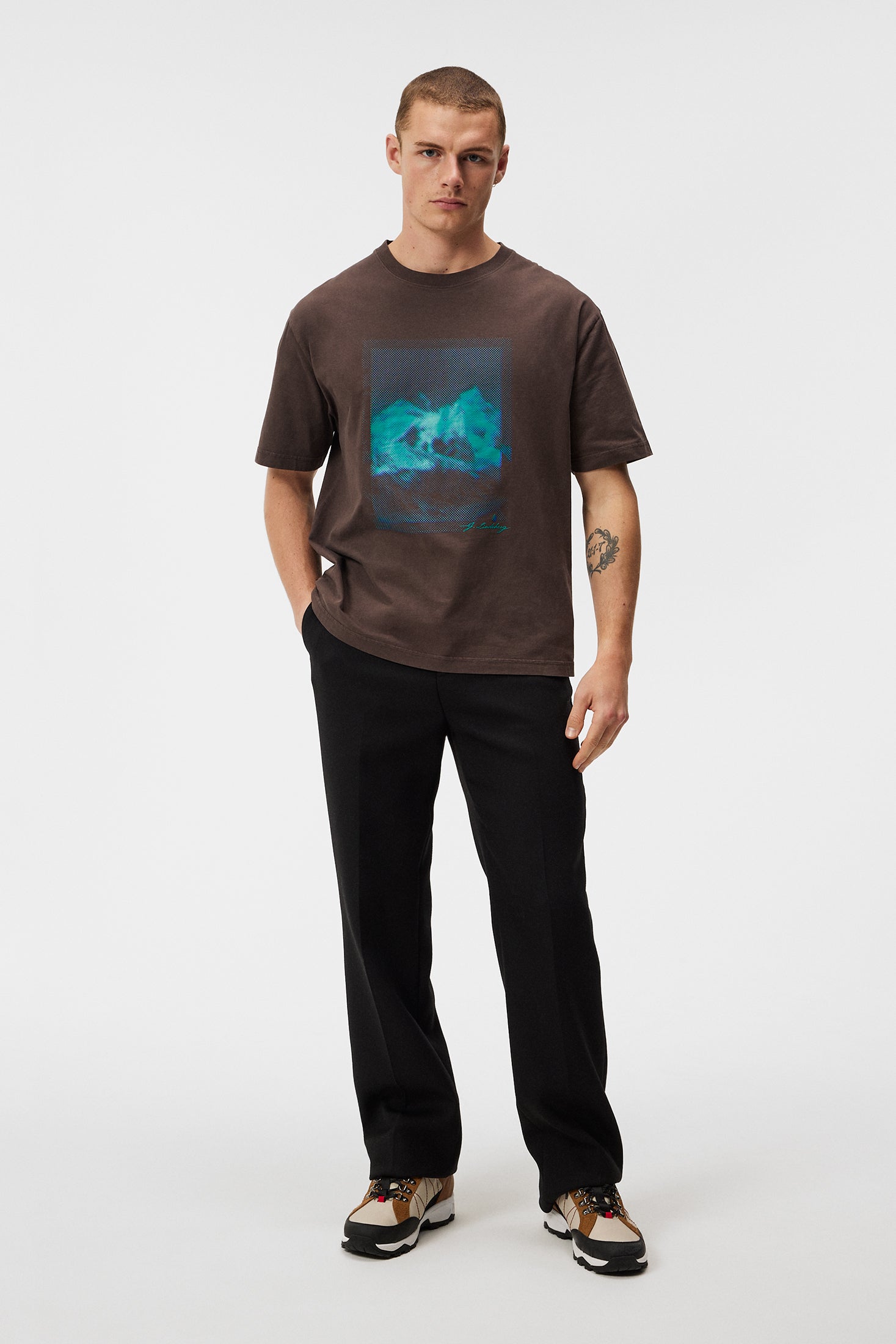 Mann mit T-Shirt und Sneakers Mann trägt braunes T-Shirt mit blauem Print, schwarze Hose und Sneakers vor weißem Hintergrund.