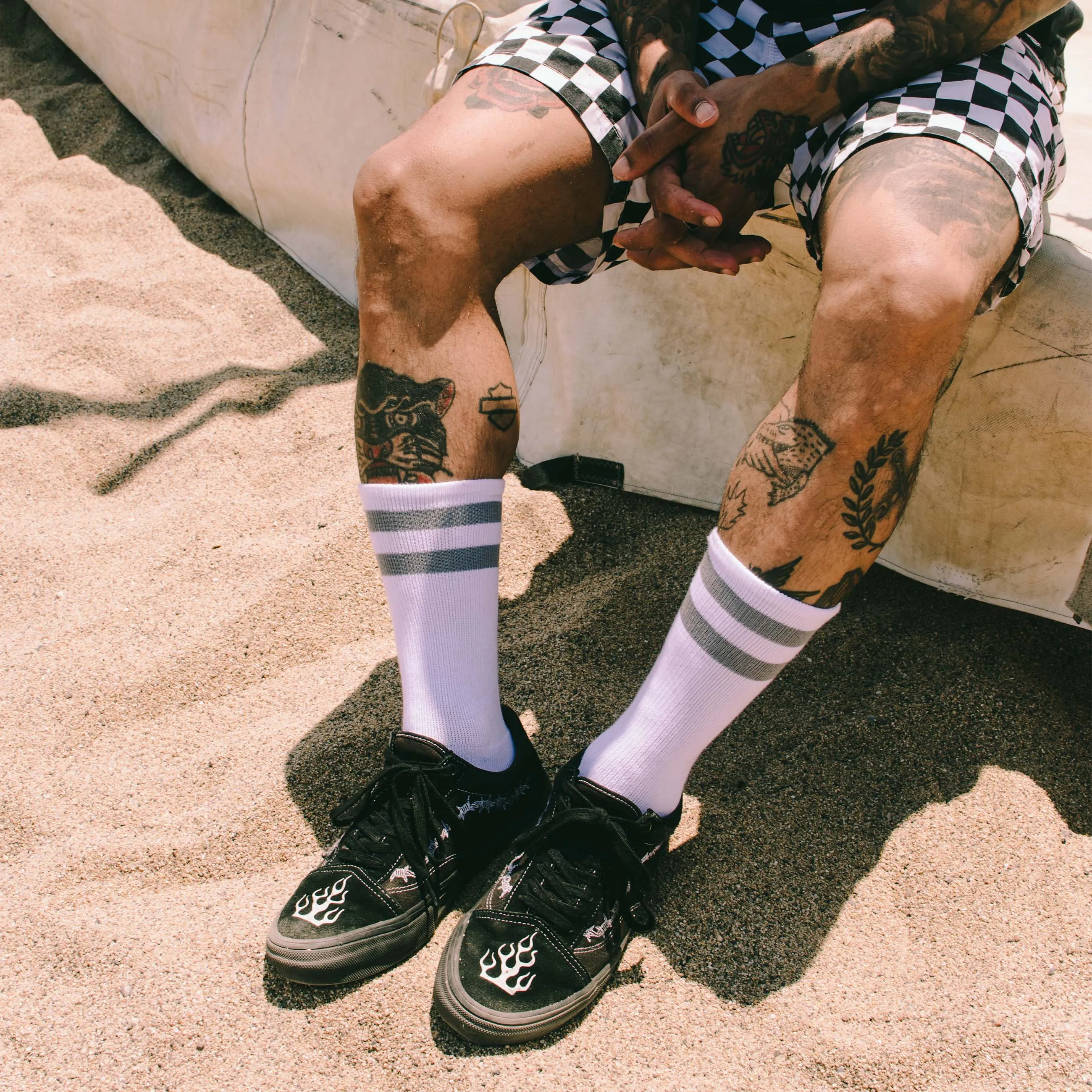 Person mit Tattoos trägt Schachbrettmuster-Shorts, Falkor Mid High Socken, und Turnschuhe am Strand.