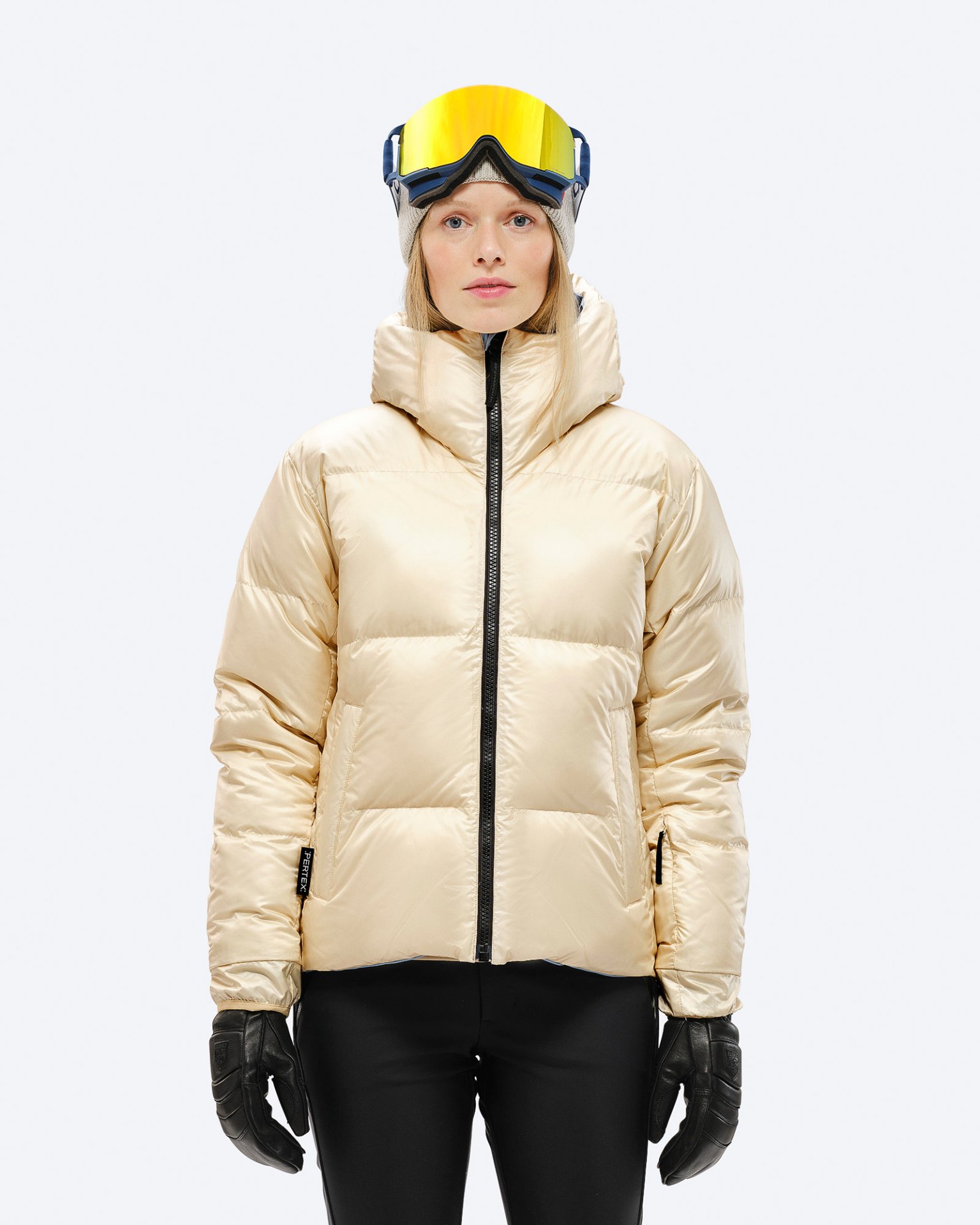 Eine Frau trägt eine glänzende, beige Daunenjacke mit Kapuze, schwarze Handschuhe und Skibrille vor einem weißen Hintergrund.