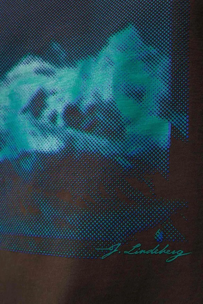 Detailaufnahme des Walsh Boxy Print Tee Delicioso mit blau-grünem, saisonalem Print und J. Lindeberg Schriftzug auf braunem Stoff.