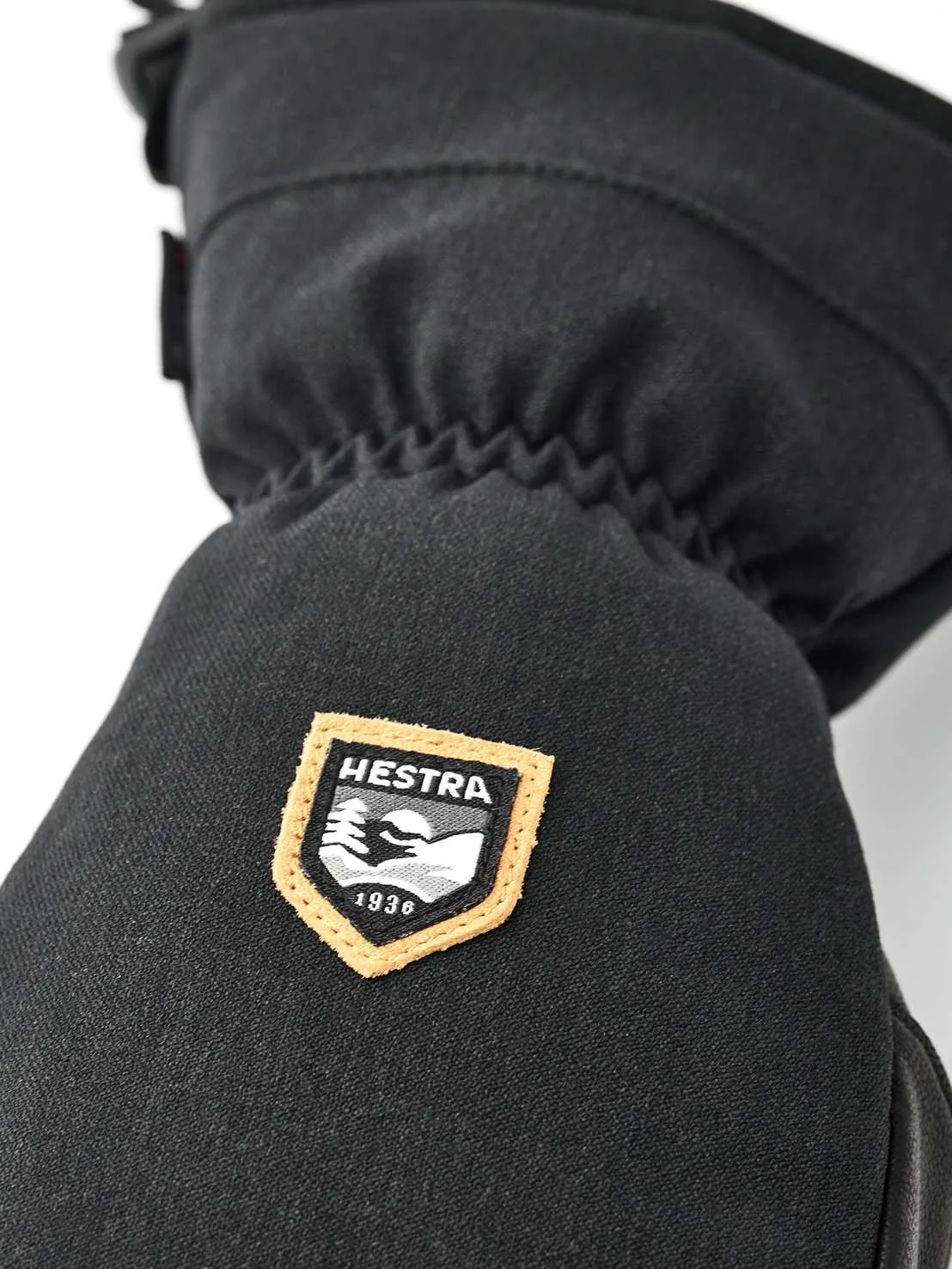 Detailaufnahme eines schwarzen Fäustlings mit dem Hestra-Logo, einem orange umrandeten Schild mit Bergmotiv und dem Gründungsjahr 1936.