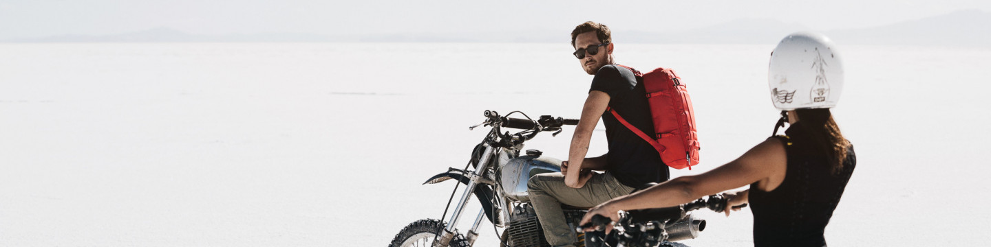 Motorradfahren mit Stil Motorradfahrer mit rotem Rucksack und Sonnenbrille sitzt auf Maschine. Frau mit Helm hält das Motorrad fest. Weißer Hintergrund.