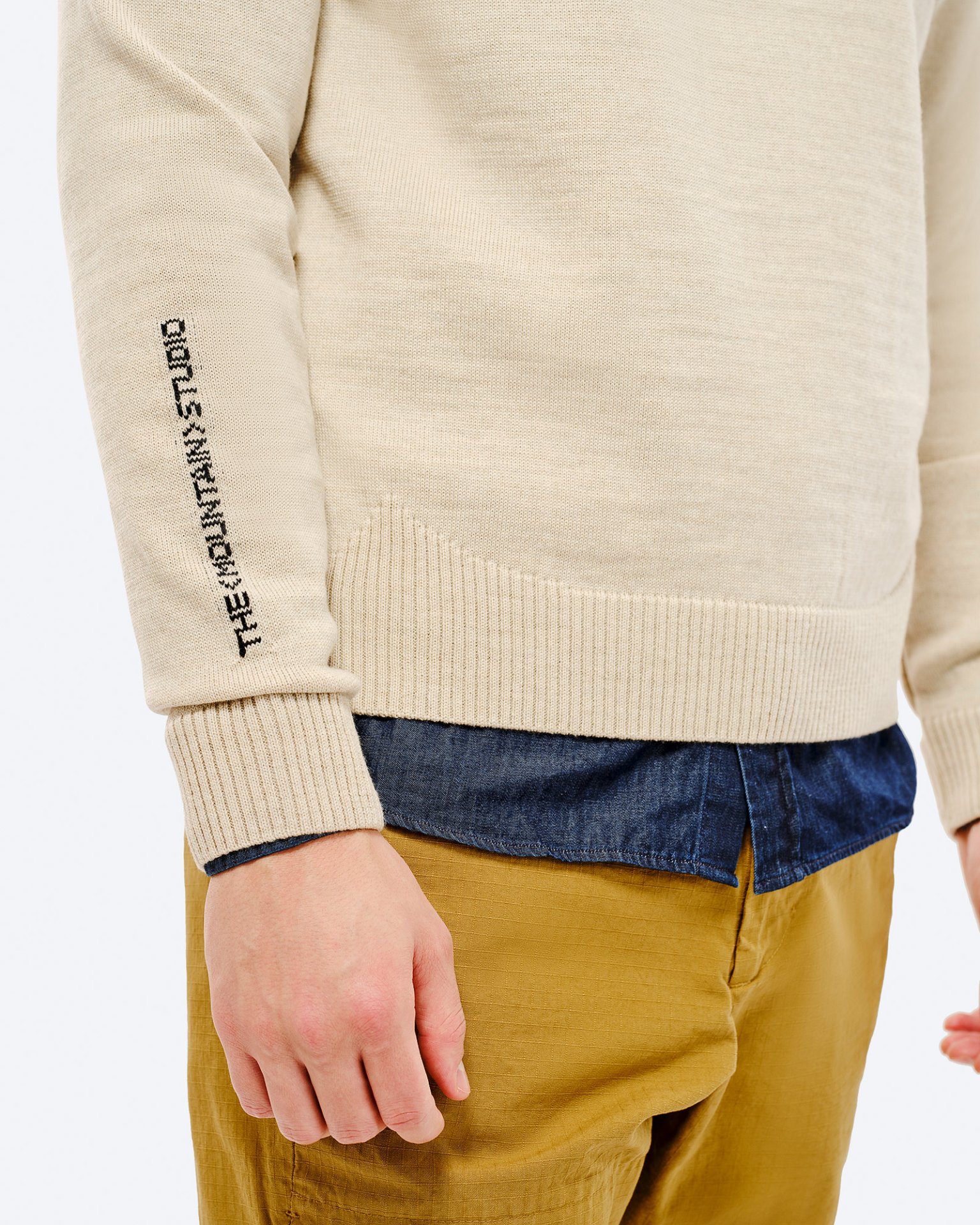 Detailansicht vom St. Moritz Ski Sweater, beige, mit Schriftzug auf dem Ärmel, über einem blauen Hemd und beigen Hosen.
