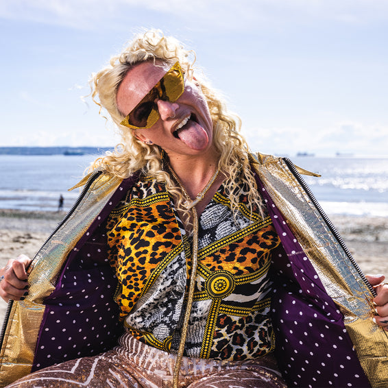 Blonde Person mit Sonnenbrille, Zunge raus, trägt eine Jacke mit Pyrit-farbenen Akzenten.