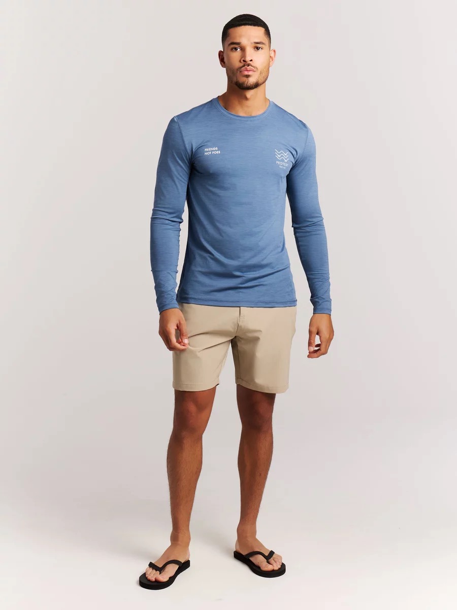 Mann in blauem Langarmshirt, beigen Shorts und Zehentrennern vor weißem Hintergrund.