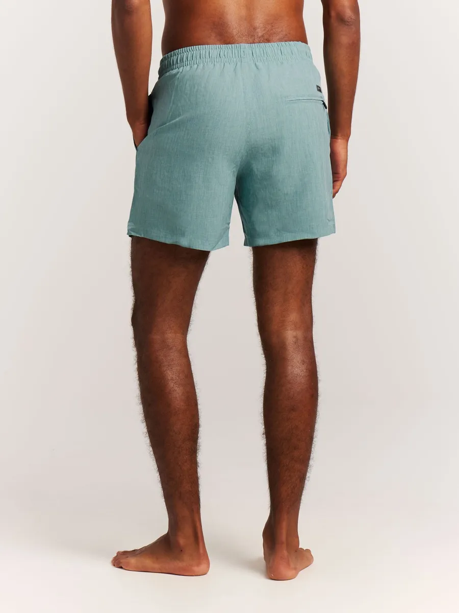 Eine Person von hinten, barfuß, mit türkisfarbenen Shorts vor weißem Hintergrund.