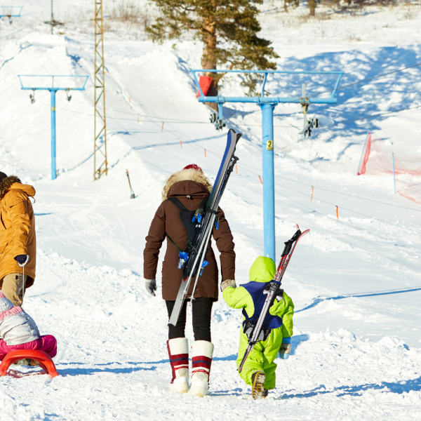 Winterliche Szene: Familie transportiert Skier mit Skiback-System auf schneebedeckter Piste. Kind trägt blaue Skier.
