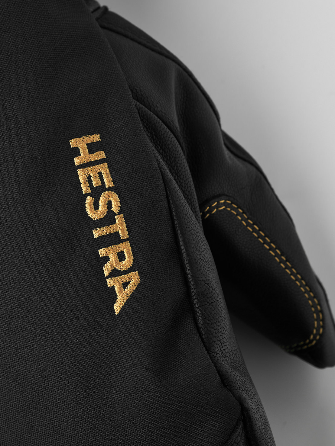 Detailansicht eines schwarzen Hestra-Handschuhs mit goldener 'HESTRA'-Stickerei und gelben Nähten.