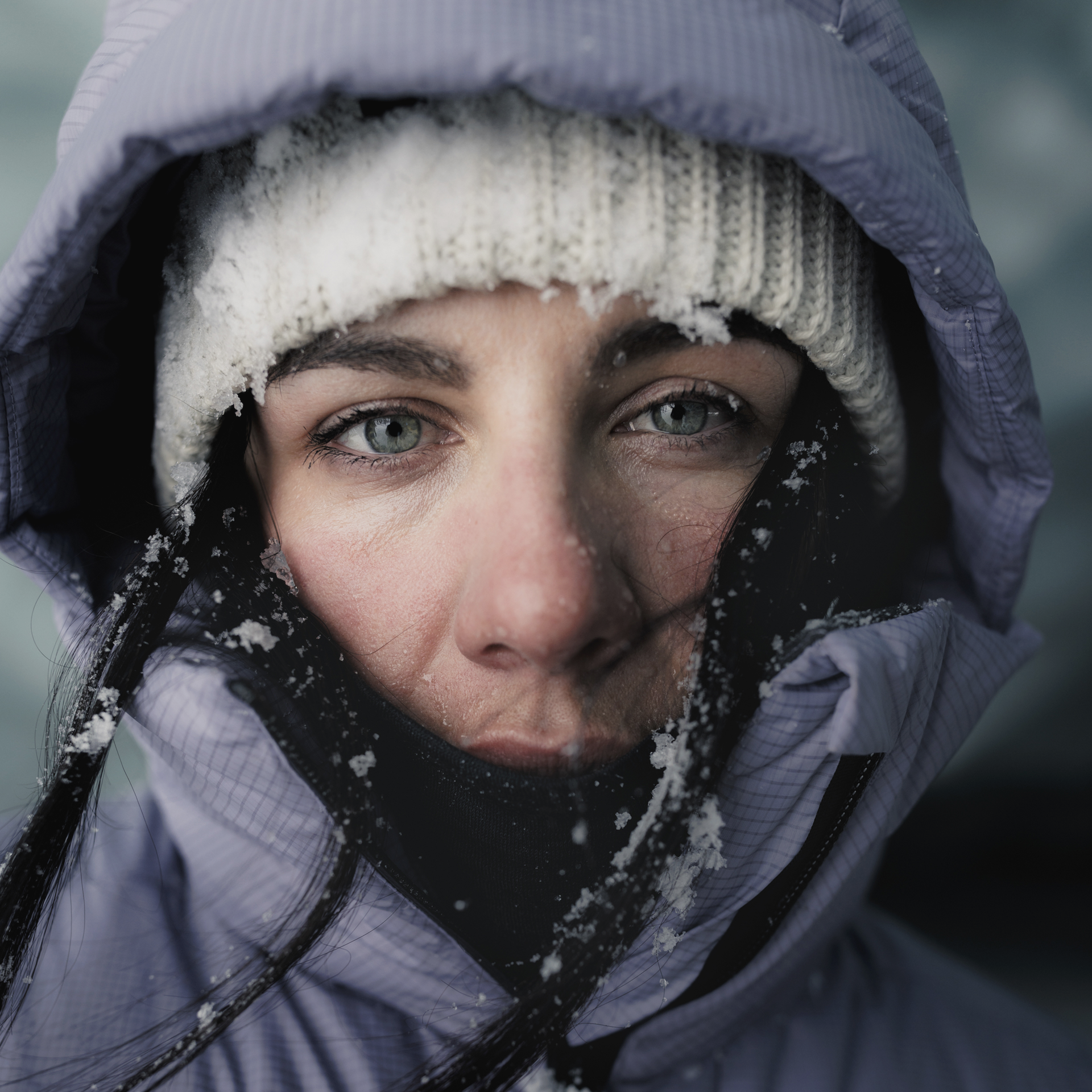 Frau mit eisbedeckter Mütze Nahaufnahme einer Frau mit eisbedeckter Mütze und Kapuze, die direkt in die Kamera blickt. Sie hat grüne Augen und dunkle Haare.