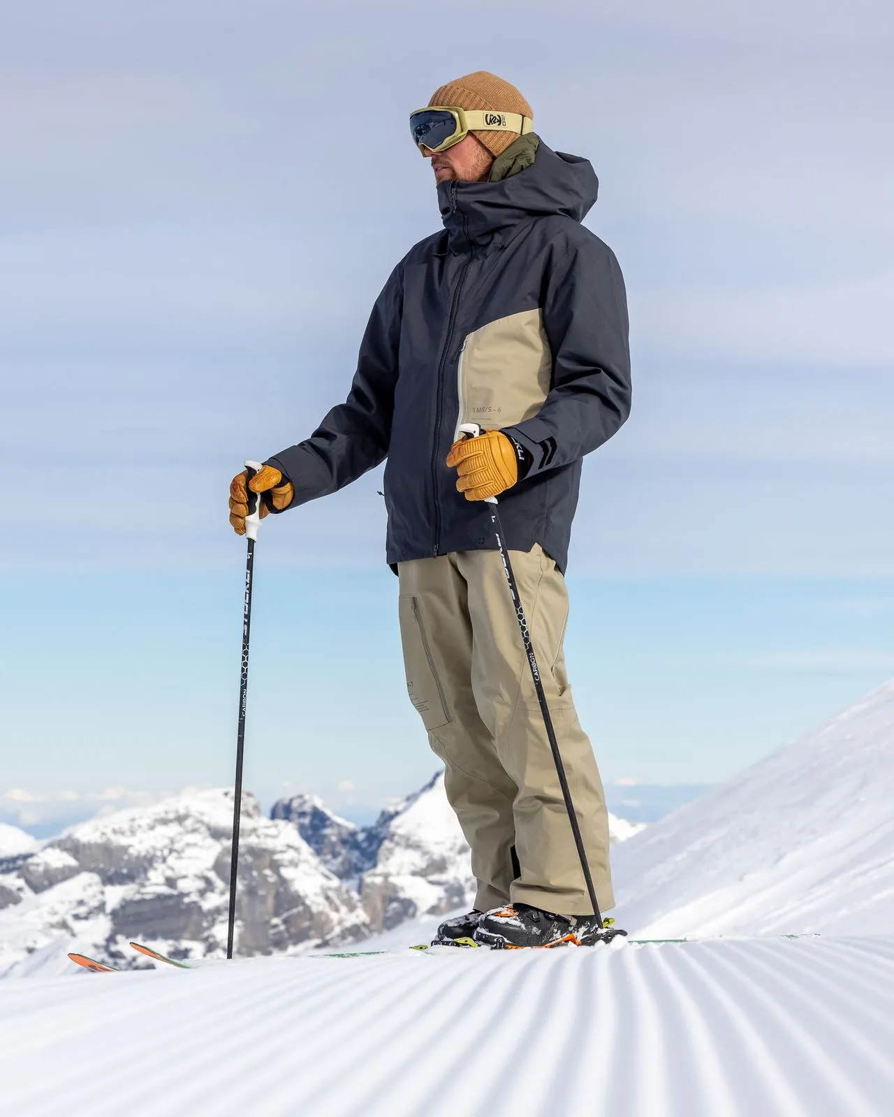 Skifahrer in P-7 Ride Insulated Pant Seneca Rock, mit Mütze, Brille und Skistöcken auf präparierter Piste.