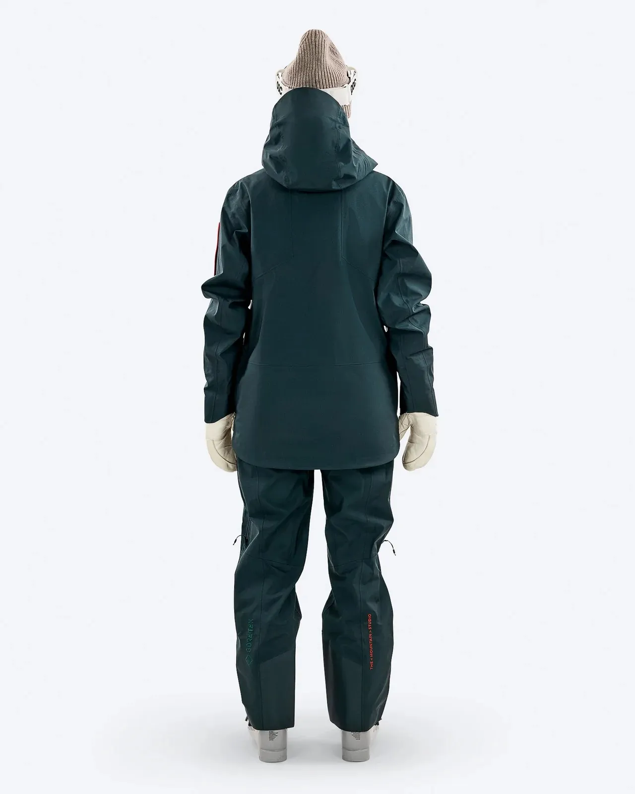 Rückansicht einer Person in dunkelgrüner Skijacke und Hose, mit Mütze und Handschuhen vor weißem Hintergrund.