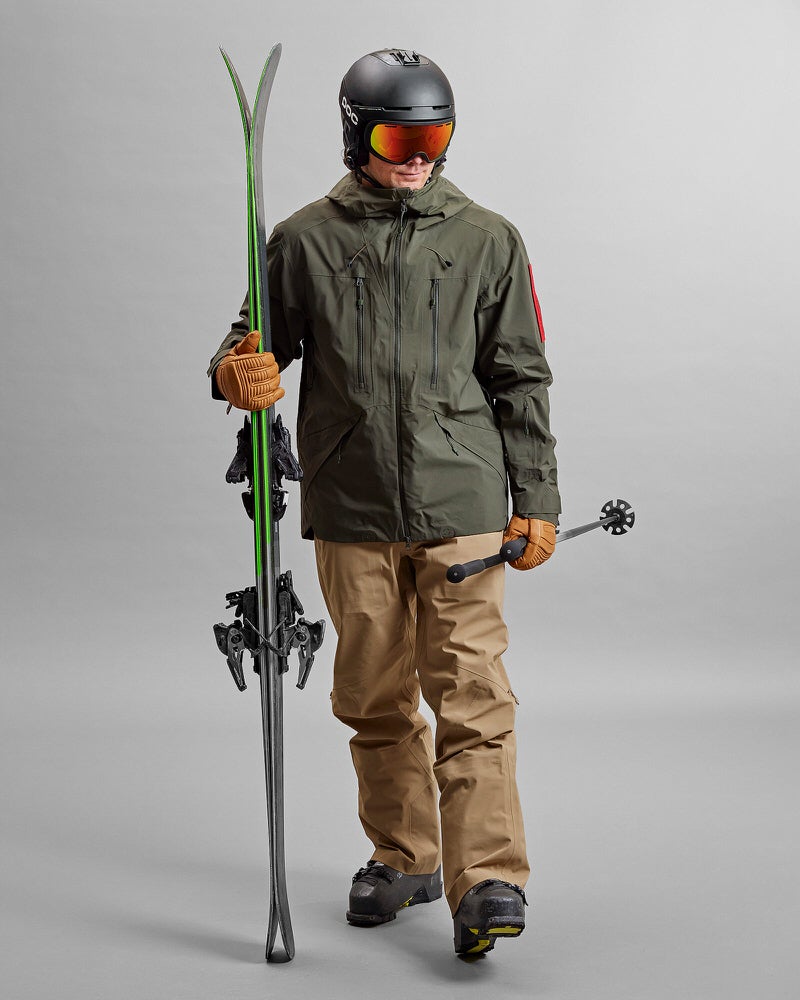 Skifahrer mit Z-2 HD GORE-TEX Pro 3L Shell Jacket Forest Green, Ski, Stöcken und Helm vor grauem Hintergrund.