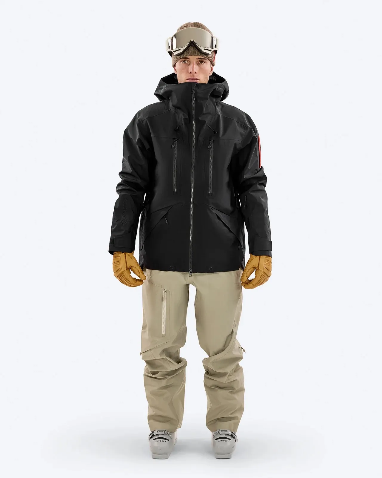 Mann in schwarzer Z-2 EPE Jacke, beigen Hosen, Handschuhen und Skibrille vor neutralem Hintergrund.