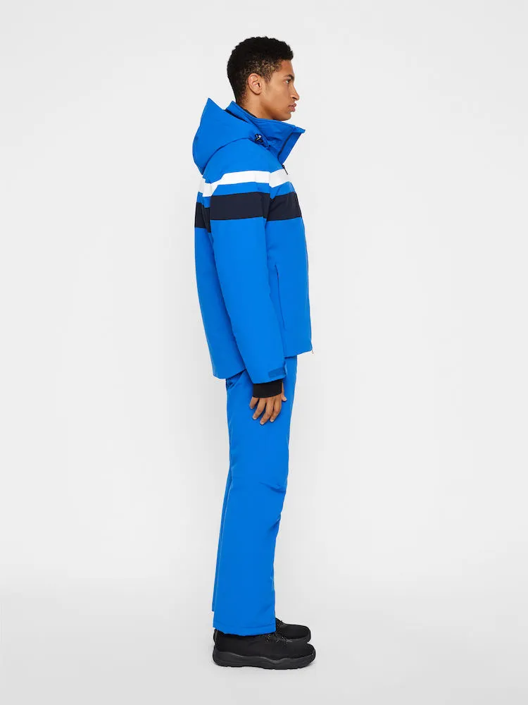 Ein Mann trägt eine blaue J.Lindeberg Franklin 2L Skijacke und Hose im Profil. Die Jacke hat weiße und marineblaue Streifen.