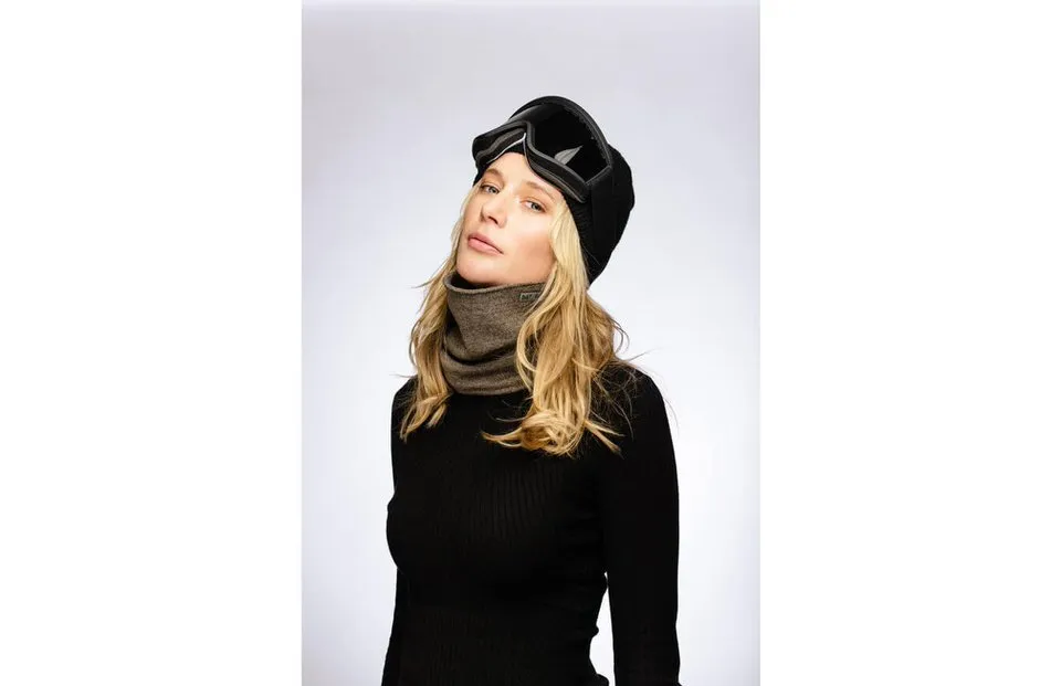 Eine Frau mit Skibrille, schwarzer Mütze und beigem Drifter Merino Neckwarmer in Kaki vor hellem Hintergrund.