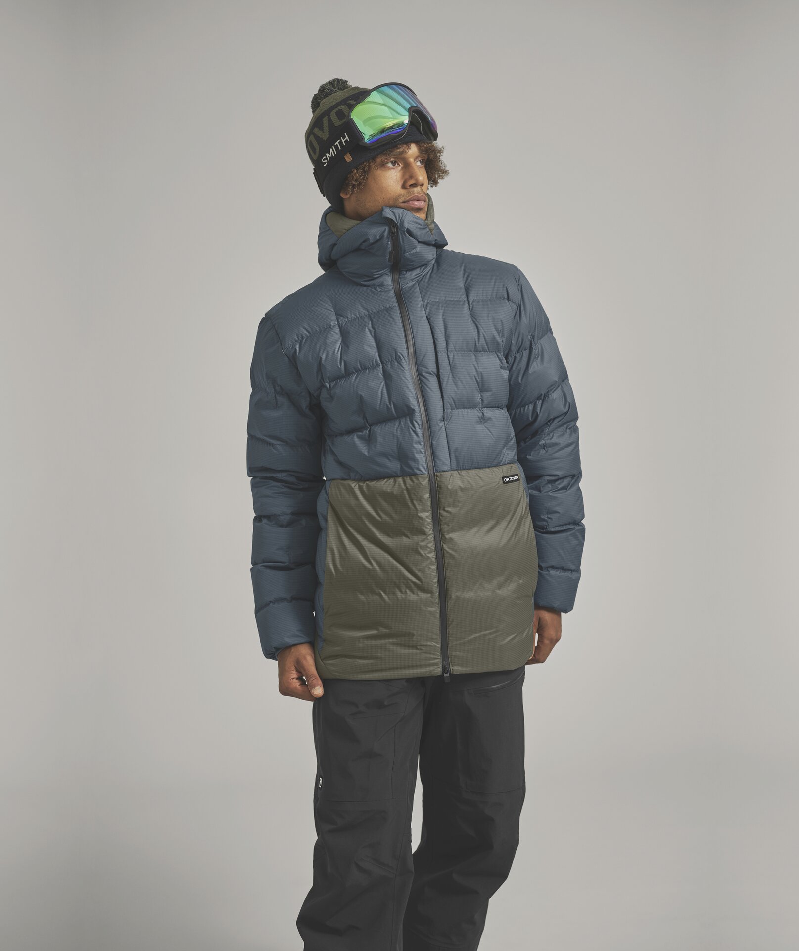 Skifahrer im Winterlook Ein Mann mit Skibrille und Mütze trägt eine zweifarbige Daunenjacke in Blau und Grün und schwarze Skihosen.