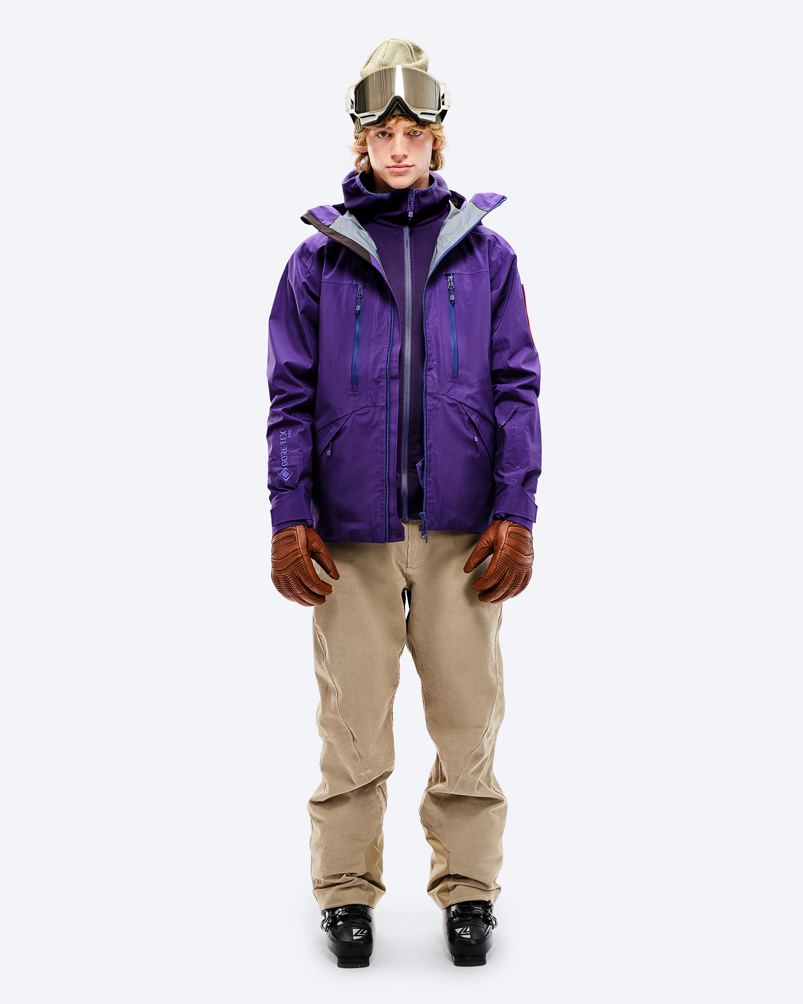 Mann in violetter GORE-TEX EPE 3L Shell Jacket, beigen Hosen, Skischuhen und Brille, frontal auf Weiß.